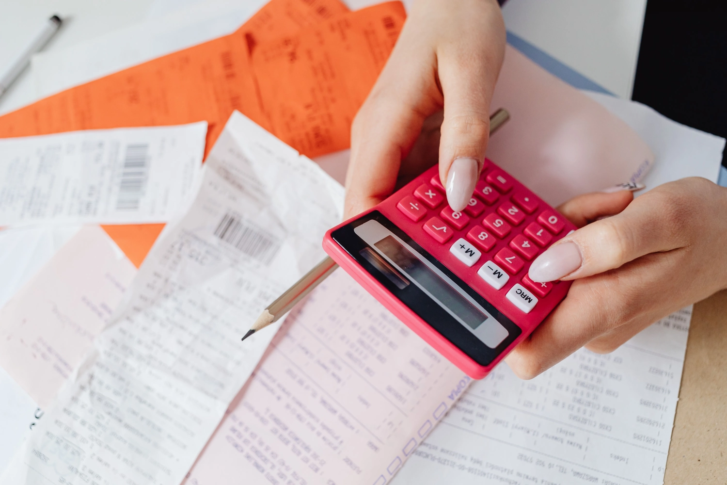 Pay Solution – Lohn- und Gehaltsberatung – Das Bild zeigt zwei Hände, die mit einem leuchtend pinken Taschenrechner Ausgaben zusammenrechnen. Um sie herum liegen verstreute Papierbelege und ein Bleistift auf einem Schreibtisch.