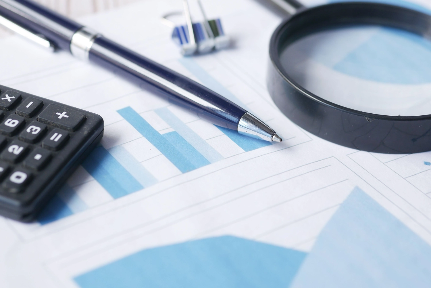 Pay Solution – Gehaltsabrechnungs- und Gehaltsberatung – ein Finanzaudit- oder Datenanalyse-Setup mit einem blauen Stift, einer schwarzen Lupe, einem Taschenrechner und einer Heftklammer, die auf gedruckten Balkendiagrammen und Finanzberichtsdiagrammen liegt